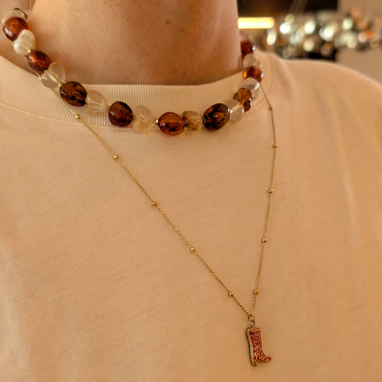 Amberkleurige kralenketting met chunky stenen en transparante harten kralen, gecombineerd met een fijne goudkleurige ketting met cowboylaars-bedel, gedragen op een zachte beige top.