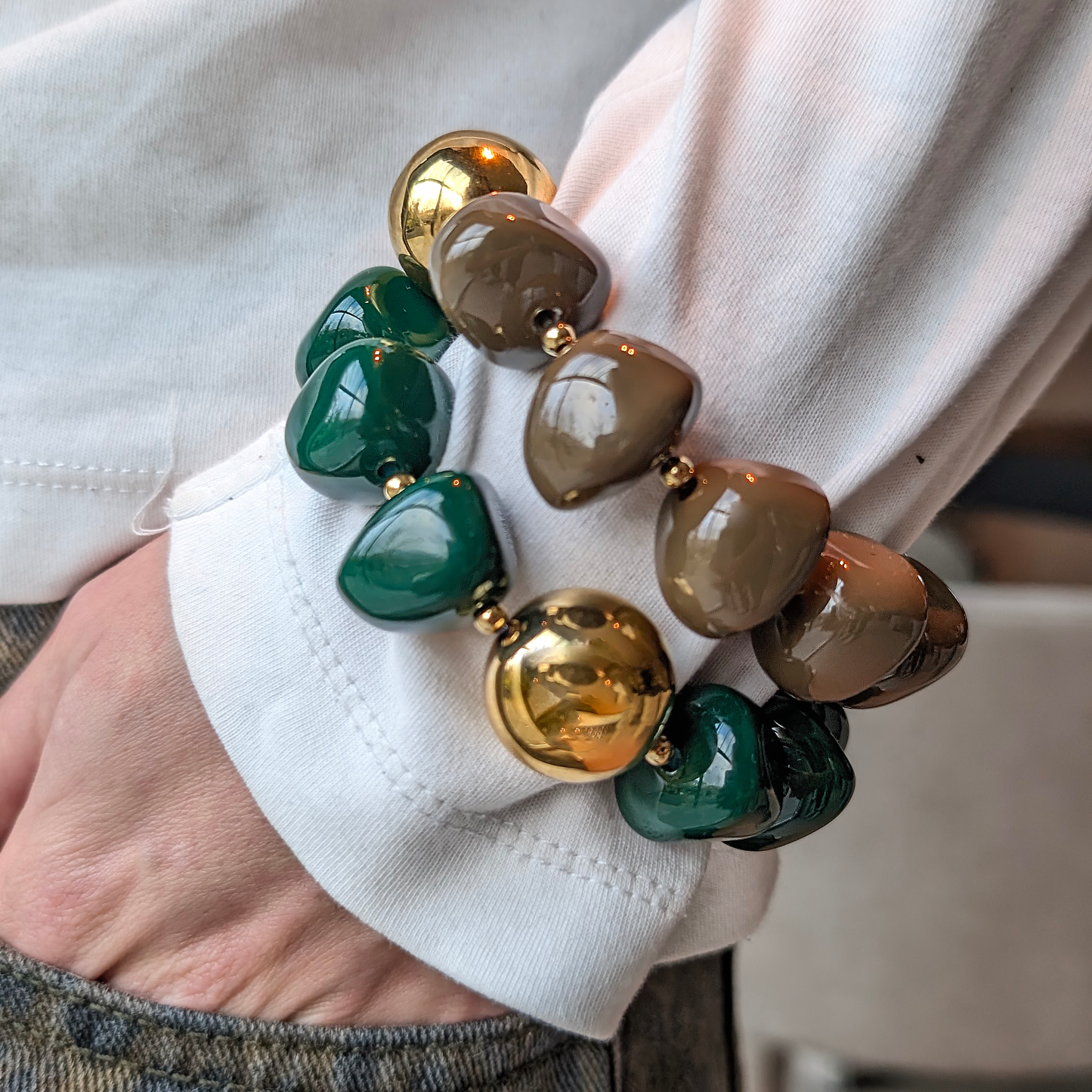 Chunky armbandenset met glanzende kralen in donkergroen en taupe, afgewisseld met grote goudkleurige parels, gedragen over de mouw van een witte top.