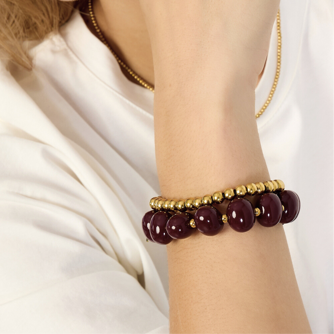 Donkerrode chunky kralenarmband met goudkleurige accenten en gouden kralenarmband gedragen om de pols bij een witte blouse.