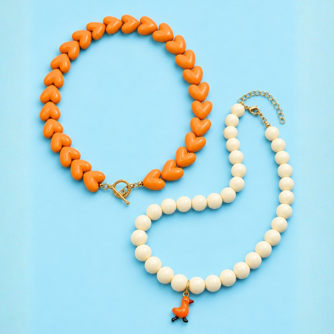 Oranje hartjesketting met goudkleurige kapittelsluiting naast een crèmekleurige kralenketting met een oranje eendenbedel.
