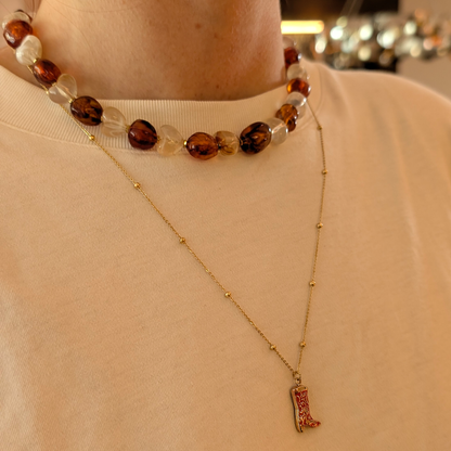Amberkleurige kralenketting  met chunky stenen en transparante harten kralen, gecombineerd met een fijne goudkleurige ketting met cowboylaars-bedel, gedragen op een zachte beige top.
