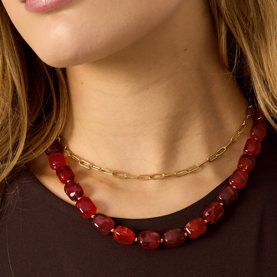 Bordeauxrode kralenketting met glanzende facetkralen, gecombineerd met een gouden schakelketting, gedragen op een donkere top.