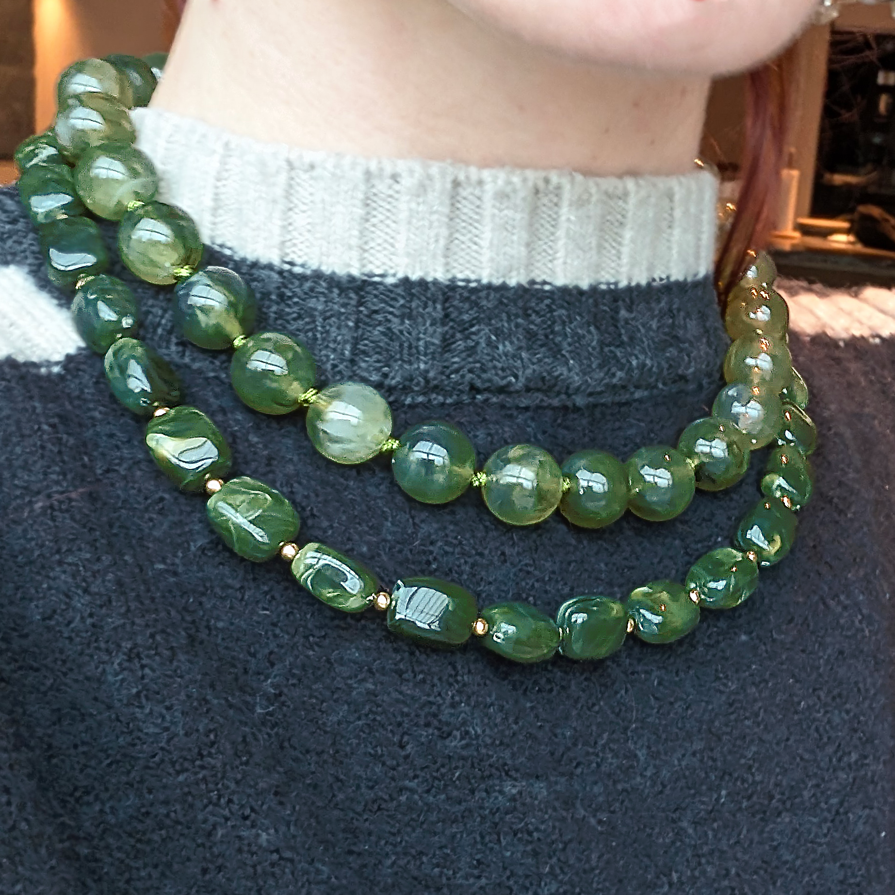 Groene kralenketting met glanzende ronde en ovale kralen en gouden tussenkralen, gedragen in een layered look op een grijs-blauwe trui.