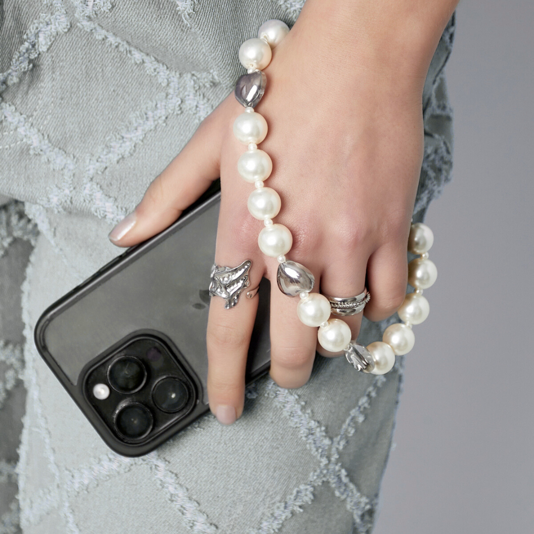 Hand met smartphone en parel telefoonkoord met zilverkleurige details, gestyled met ringen en lichte denim outfit.