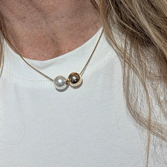 Minimalistische ketting met een goudkleurige en een parelmoer kraal, gedragen op een witte top.