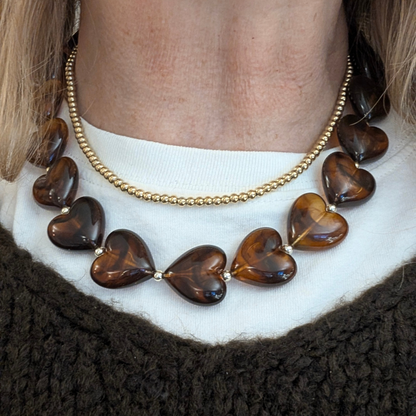 Model met wit tshirt en bruine trui met 2 kettingen met goudkleurige bolletjes en amberbruine gemarmerde hartjeskralen, gedragen op een witte top, warme Ibiza-style uitstraling.