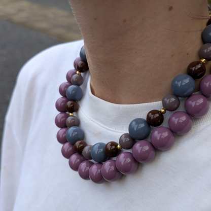 Dubbele chunky kralenketting in paars, bruin en grijsblauw – glanzende grote kralen met gouden details, gedragen op een witte top.