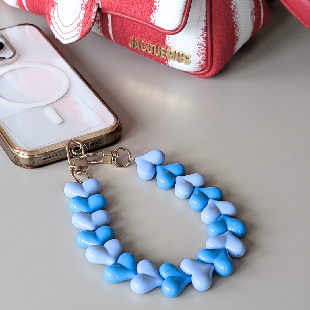 Telefoonkoord met hartvormige kralen in lichtblauw en blauw, duo hartjes design met goudkleurige clip, bevestigd aan smartphone.