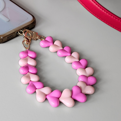 Telefoonkoord met hartvormige kralen in verschillende roze tinten, duo hartjes design met goudkleurige clip, bevestigd aan smartphone.