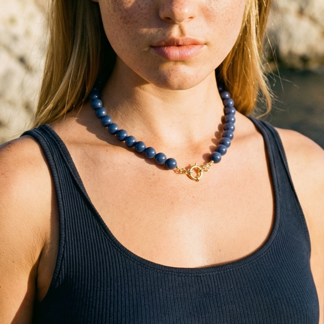 Donkerblauwe kralenketting met ronde matte kralen en goudkleurige boeisluiting, gedragen op een donkerblauwe top in een natuurlijke setting – stijlvolle statement ketting