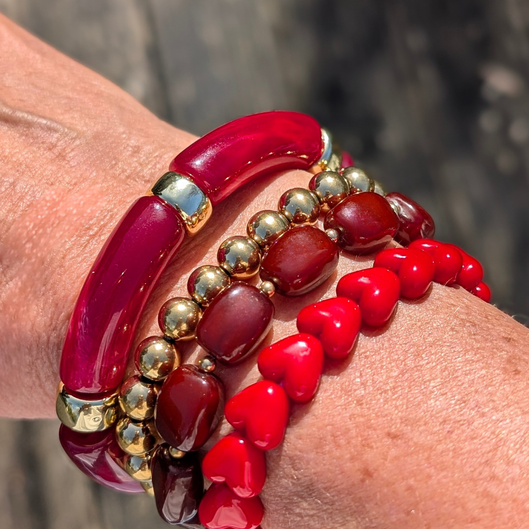 Arm met donkerrode tube armband, gouden kralen armband, bruine kralen armband en rode hartjes armband