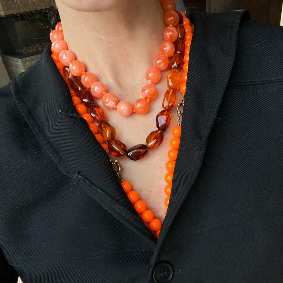 Drie lagen oranje kralenkettingen, bestaande uit feloranje ronde kralen, marmer-look perzikkleurige kralen en facetgeslepen amberbruine kralen, gedragen op een navy blazer.