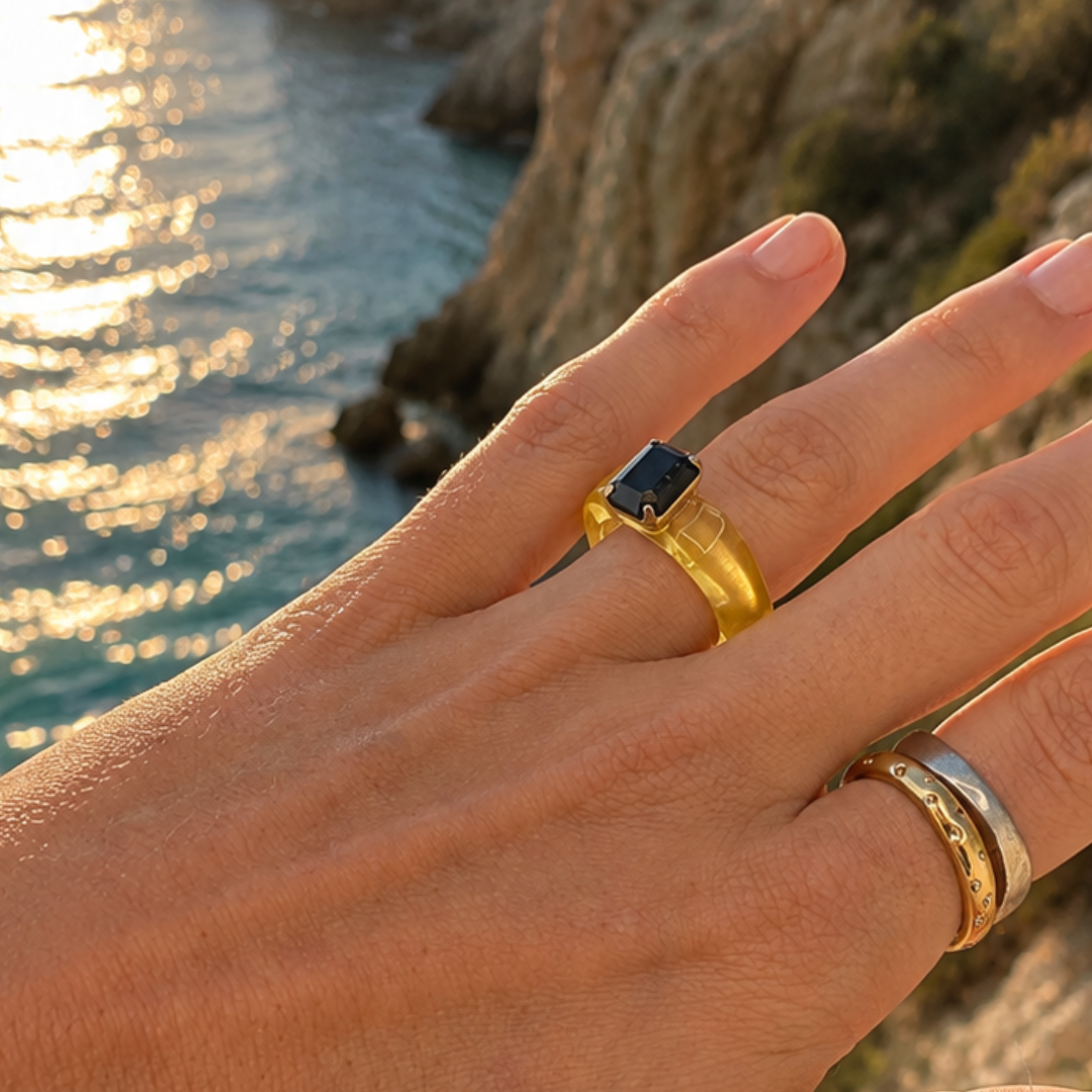 Hand met de gele chunky ring met zwarte steen en goudkleurige ringen, met zee en rotsen op de achtergrond.