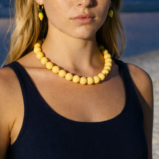 Gele kralenketting met ronde glanzende kralen, gedragen op een donkerblauwe top aan zee met citroen oorringetjes – zomerse statement ketting met Ibiza vibe