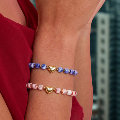 Set van twee kralenarmbanden met blauw-witte en roze-witte gestreepte kralen en een goudkleurig hartje, gedragen om de arm bij een rode jurk.