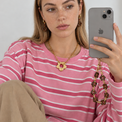 Model in roze gestreepte trui draagt de gouden bloemketting en bruine bloem oorbellen, met het bloemen telefoonkoord aan haar telefoon.