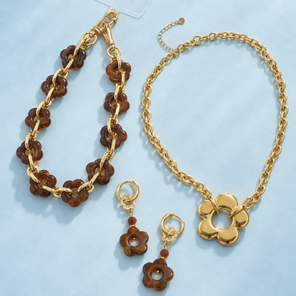 Goud-bruine bloemenset met gouden schakelketting met bloem, bruine bloem oorbellen en telefoonkoord met gemarmerde bloemen op een lichtblauwe achtergrond.