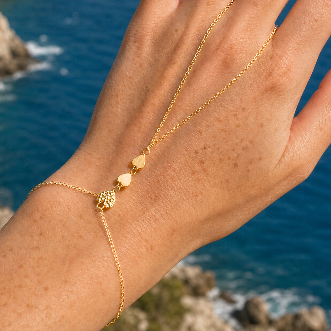 Gouden hand chain armband met hartjes en steentjes verbonden met ring en fijne schakels gedragen aan hand.