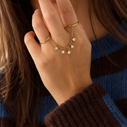 model met Gouden ring handchain met twee verbonden ringen, een fijne gouden schakelketting en vijf ronde strass bedels met heldere steentjes op een witte achtergrond.