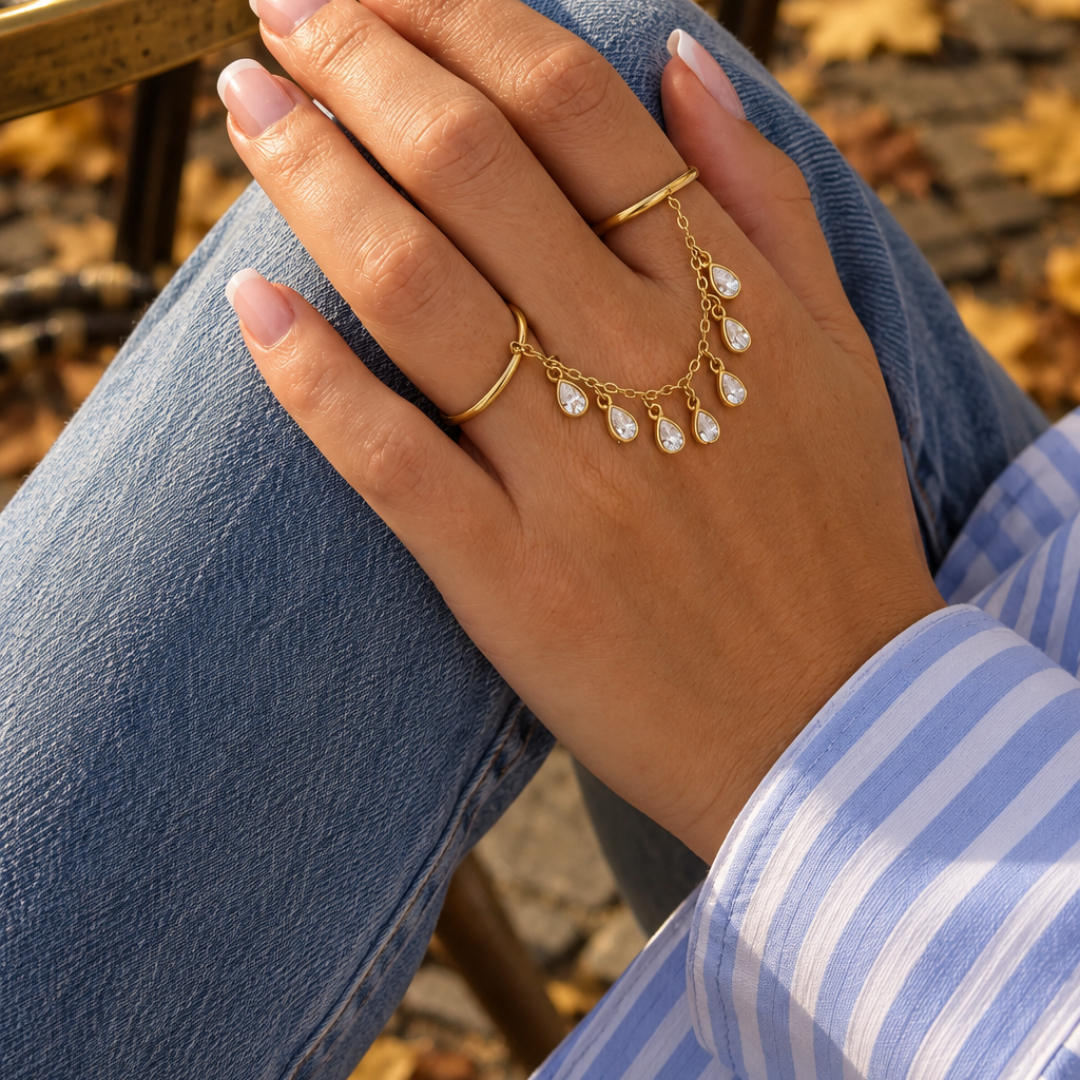 hand met Gouden ring handchain met twee verbonden ringen, een fijne gouden schakelketting en zeven hangende druppelvormige bedels met heldere strass steentjes op witte achtergrond.
