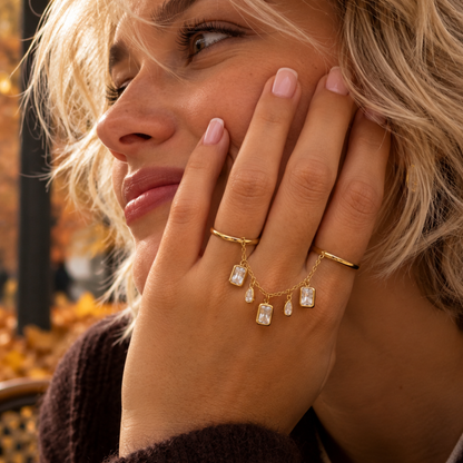model met Gouden ring handchain met twee verbonden ringen en vijf hangende strass bedels, waarvan drie rechthoekig en twee druppelvormig, op een witte achtergrond.