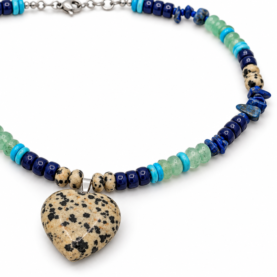 Detail van een beige harthanger met zwarte vlekjes aan een ketting met blauwe splitkralen, mintgroene kralen en turquoise accenten.
