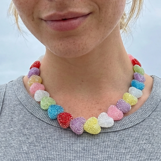Vrolijke ketting met kleurrijke suikerachtige hartvormige kralen in pastel- en felle tinten, gedragen op een grijze top – speelse statement ketting met Ibiza vibe