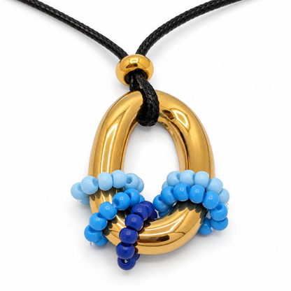 Close-up van de goudkleurige ovale ringhanger met blauwe kraaltjes rond de onderkant en een zwart gevlochten koord.