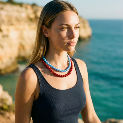 Model draagt twee Ibiza-style kralenkettingen met ronde kralen in lichtblauw en rood, gedragen in laagjes, gefotografeerd aan zee met zomerse uitstraling