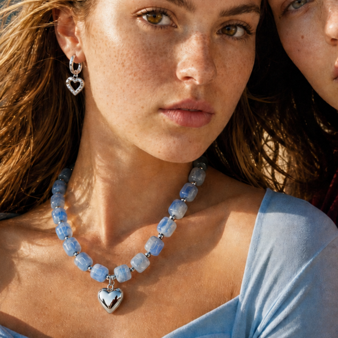 Model draagt een lichtblauwe chunky kralenketting met zilverkleurige hartbedel en zilveren hart oorbel van Casa Jadel