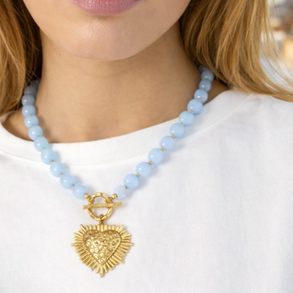 Model draagt een lichtblauwe kralenketting met gouden hart hanger in een zomerse Ibiza strandsetting – Casa Jadel