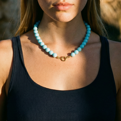 Lichtblauwe kralenketting met ronde matte kralen en goudkleurige sluiting, gedragen op een donkerblauwe top in een natuurlijke setting – frisse statement ketting