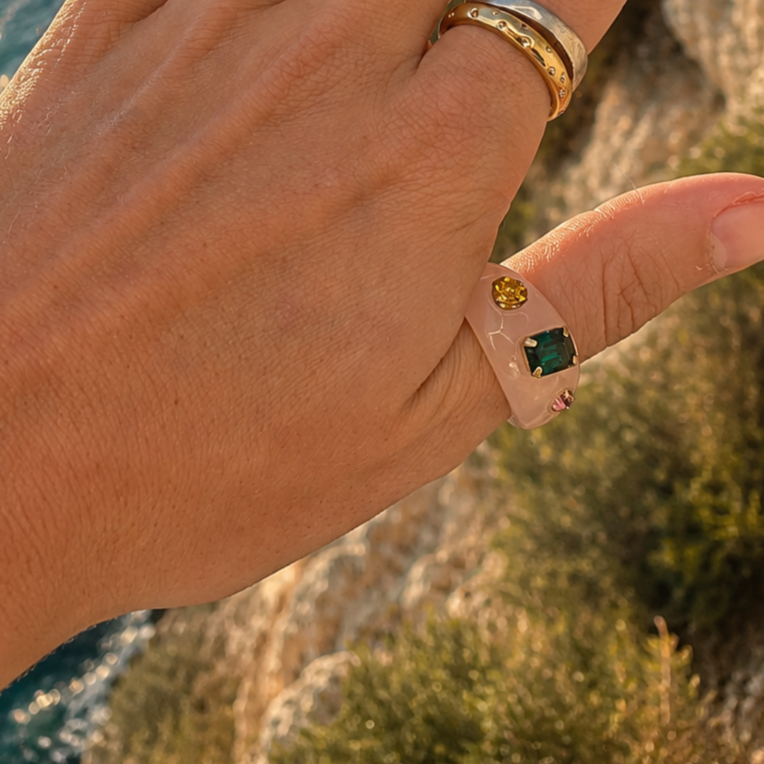 Hand met de lichtroze chunky ring met gekleurde steentjes om de duim, met zee en rotsen op de achtergrond.