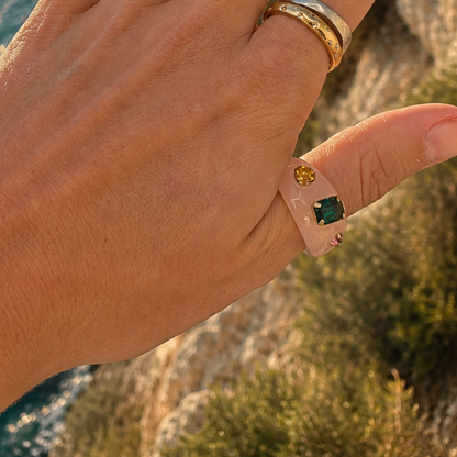 Hand met de lichtroze chunky ring met gekleurde steentjes om de duim, met zee en rotsen op de achtergrond.