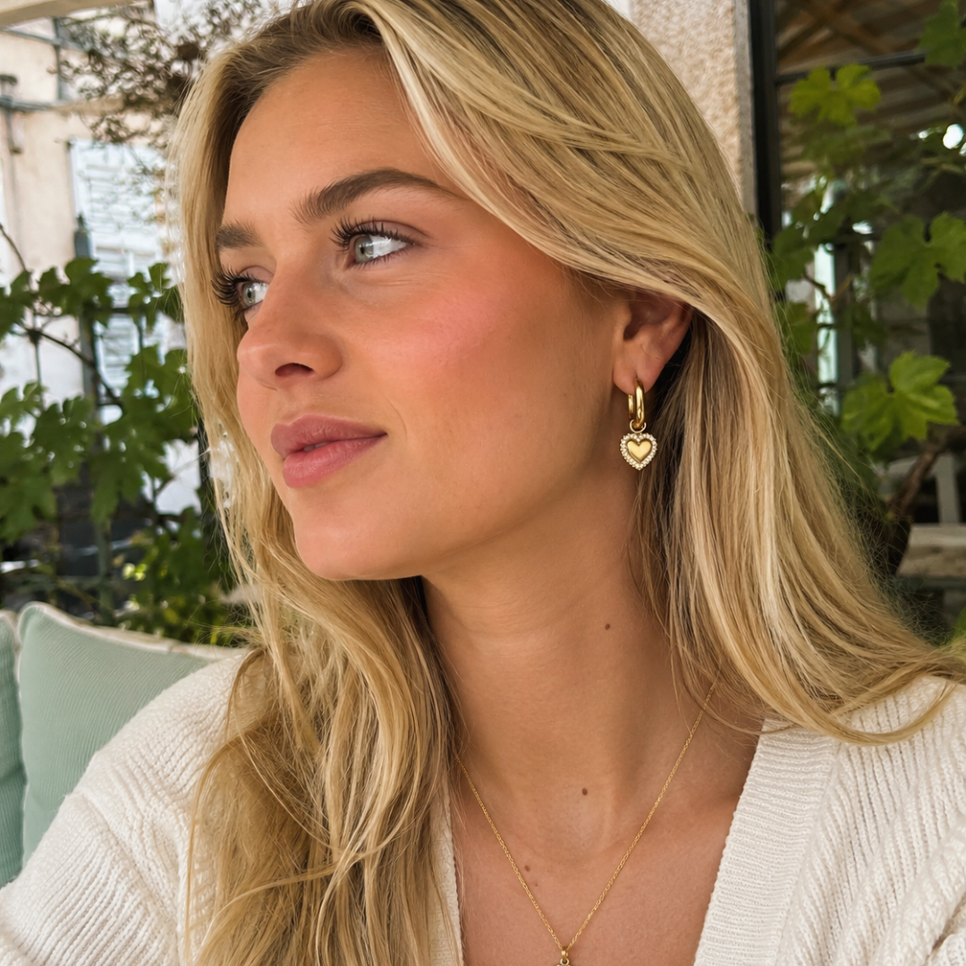 Model met Gouden ovale oorbellen met hartvormige hanger en fonkelende strass steentjes op witte achtergrond