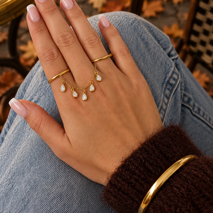 hand met Gouden ring handchain met twee verbonden ringen, een fijne gouden schakelketting en vijf hangende druppelvormige strass bedels met heldere steentjes op witte achtergrond.