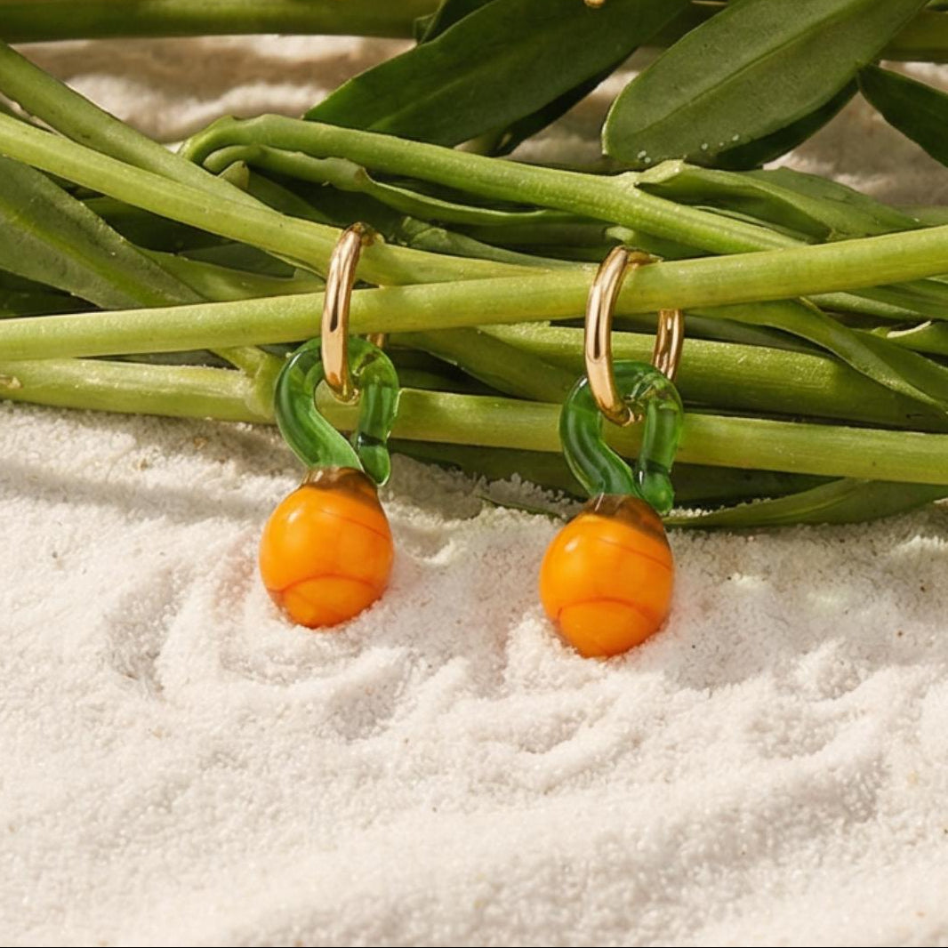 Oranje fruit oorbellen met glaskralen en gouden hoops op zand met groene takken Casa Jadel