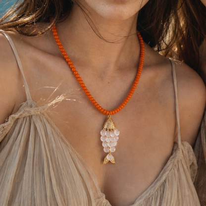 Model draagt een oranje kralenketting met roze schelpenhanger en goudkleurige visstaart van Casa Jadel
