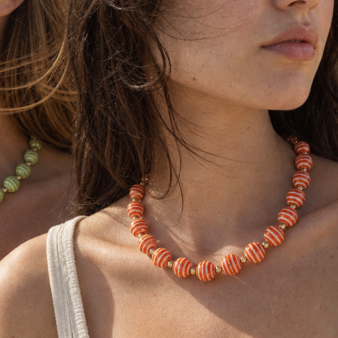 Model draagt een oranje kralenketting met wit gestreepte kralen en goudkleurige details van Casa Jadel