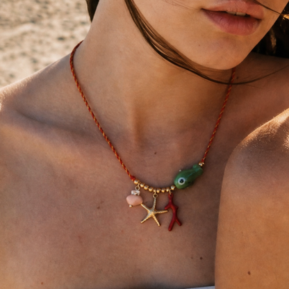 Model draagt een rode koordketting met groene vis, goudkleurige zeester, rode koraal en roze schelp bedel van Casa Jadel