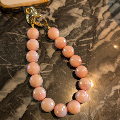 Zalmroze facetgeslepen kralen telefoonkoord met glanzende ronde disco kralen en goudkleurige sluiting, gefotografeerd op een donkere marmeren ondergrond.