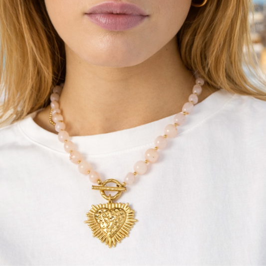 Model draagt een lichtroze kralenketting met gouden hart hanger, zomerse Ibiza uitstraling op het strand