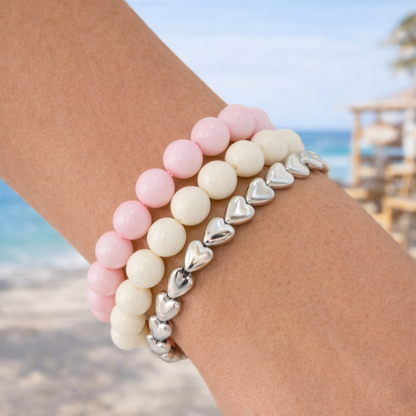 Arm van een vrouw met drie armbanden: roze kralenarmband, crème kralenarmband en zilverkleurige hartjesarmband in een zomerse strandsetting – Casa Jadel