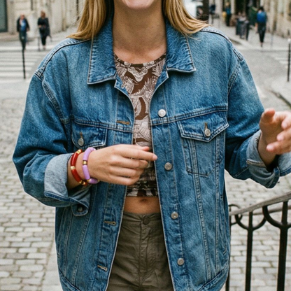model draagt meerdere kleurrijke tube armbanden, waaronder rode en lila chunky armbanden met goudkleurige details, gecombineerd met een denim jack in een stedelijke setting.