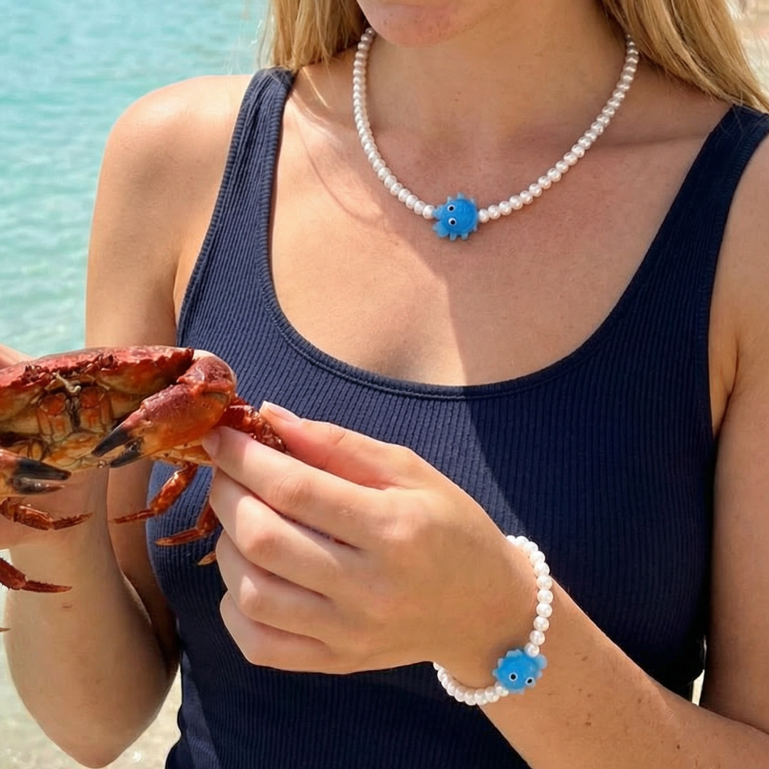 Model draagt een witte kralenketting en armband met blauwe krab bedel, zomerse sieradenset gedragen aan zee.
