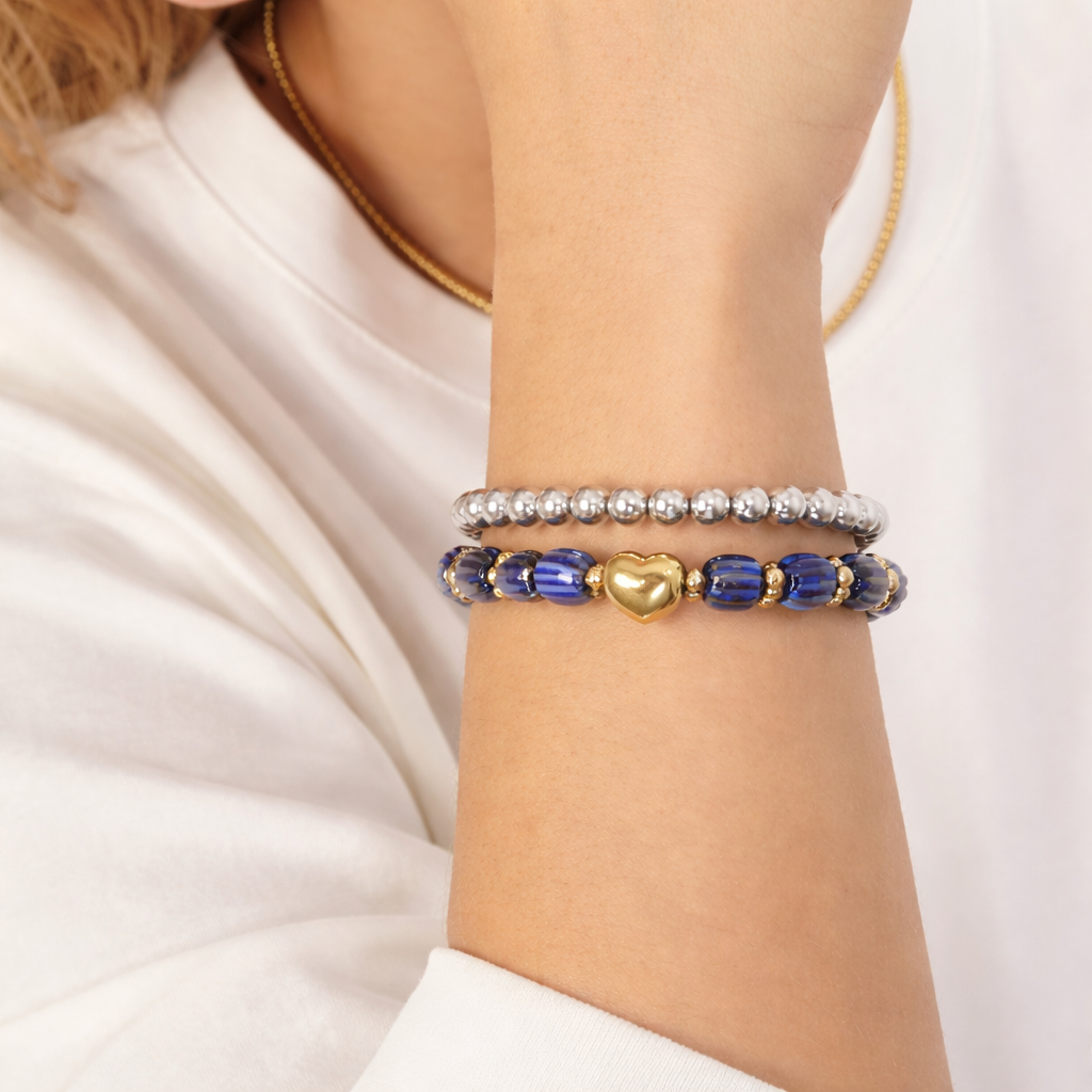 Zilveren bolletjes armband gecombineerd met blauwe kralenarmband met goudkleurig hartje, gedragen om de pols bij een witte blouse.