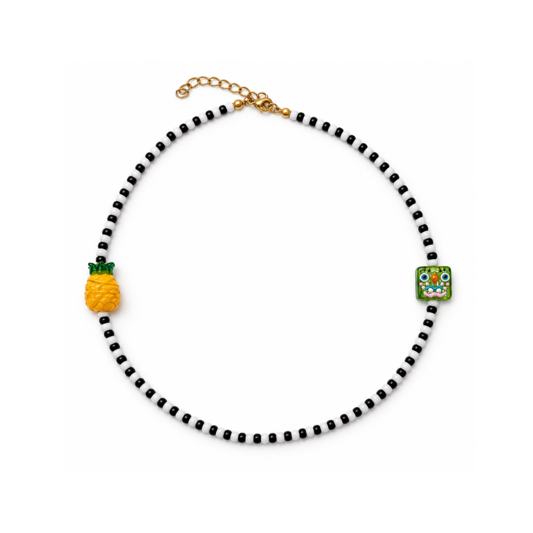 Zwart-witte kralenketting met een gele glazen ananas, groene vierkante gezichtskraal en goudkleurige sluiting met verlengkettinkje.
